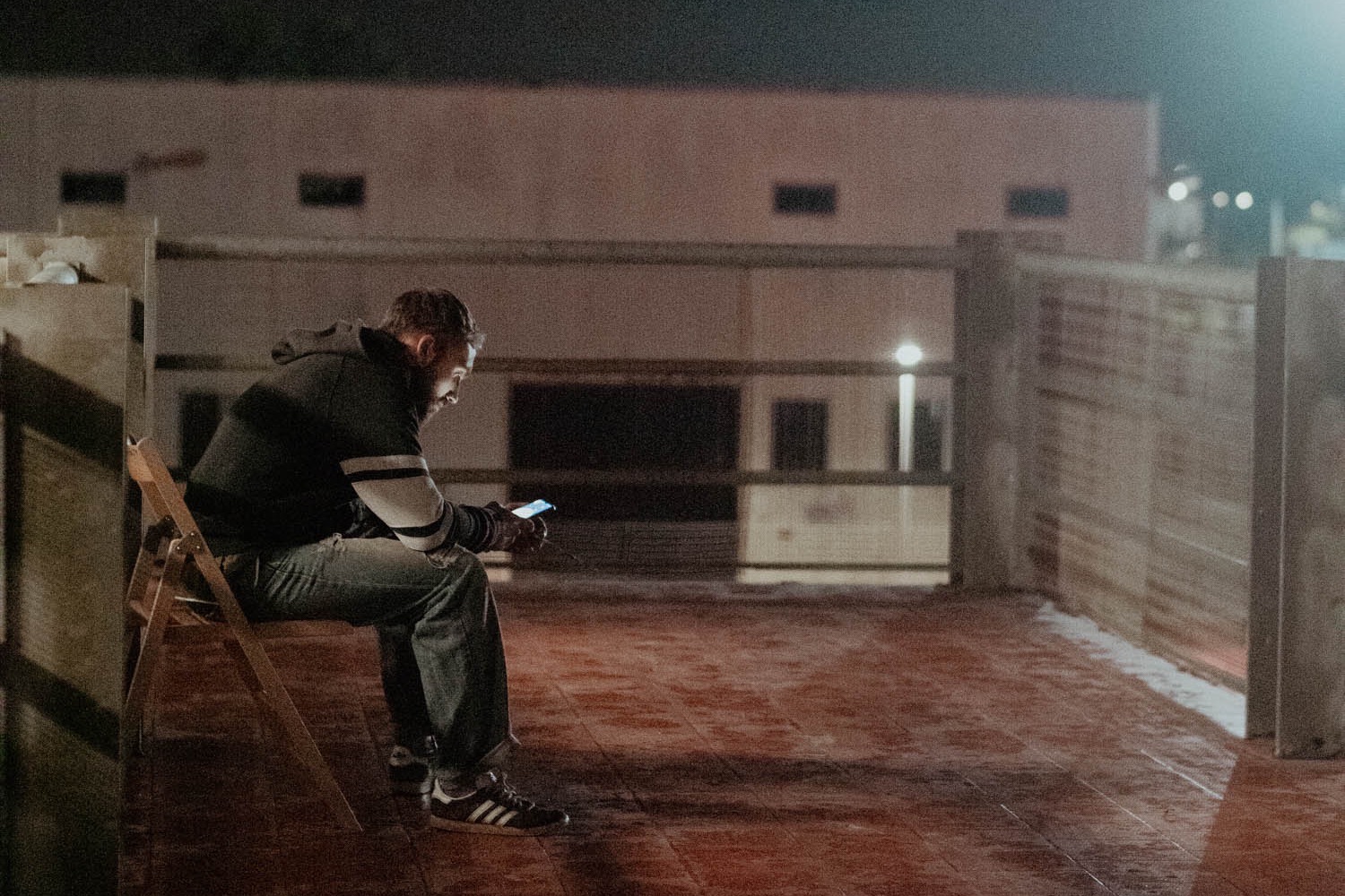 Un ospite seduto su una sedia pieghevole guarda il telefono, di notte, sul piazzale del dormitorio